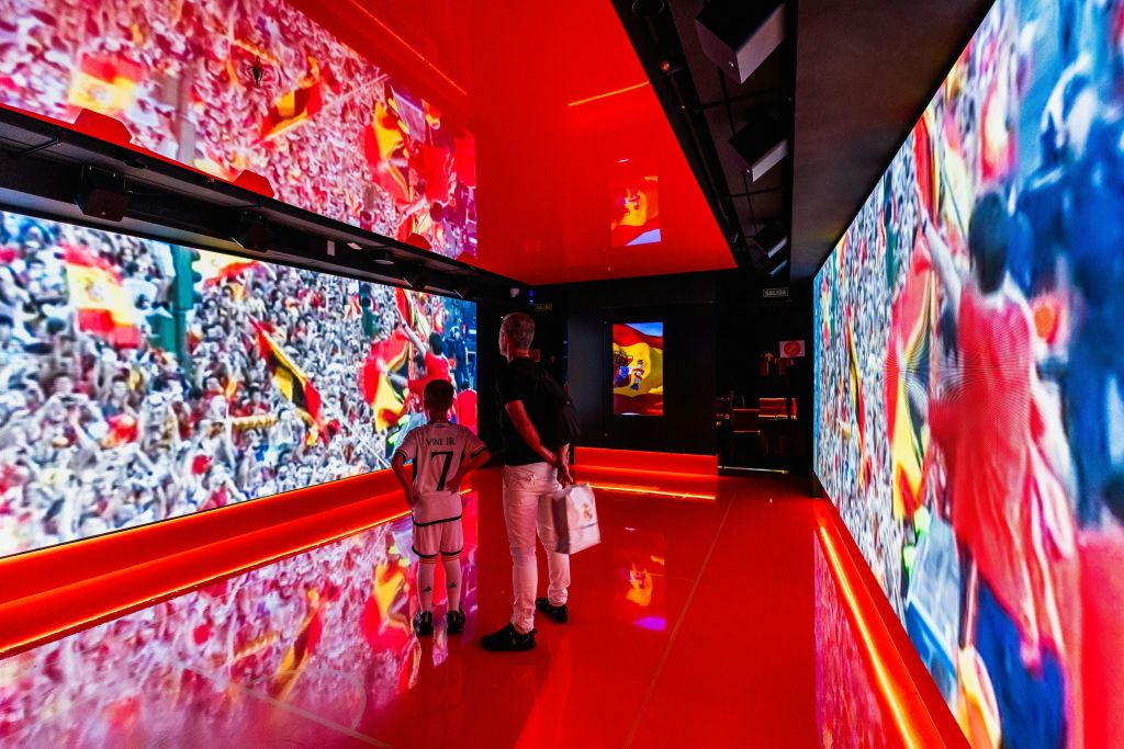 스포츠 장면과 군중 이미지를 보여주는 대형 스크린이 있는 밝고 빨간색의 방에 두 사람이 서 있습니다.