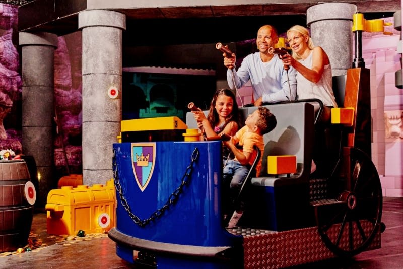 4인 가족이 방패 엠블럼이 달린 파란색 카트에서 장난감 총을 조준하며 라이딩을 즐깁니다.