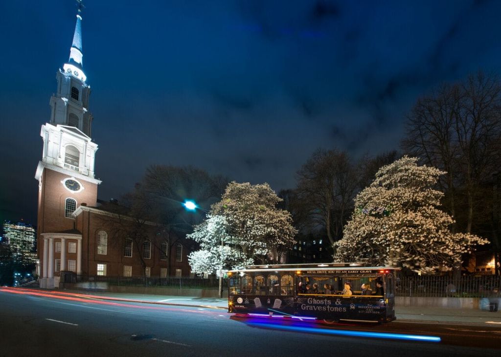 '유령과 묘비' 라는 이름의 투어 버스가 야간에 뾰족한 불빛과 꽃이 만발한 나무가 있는 교회를 지나갑니다.