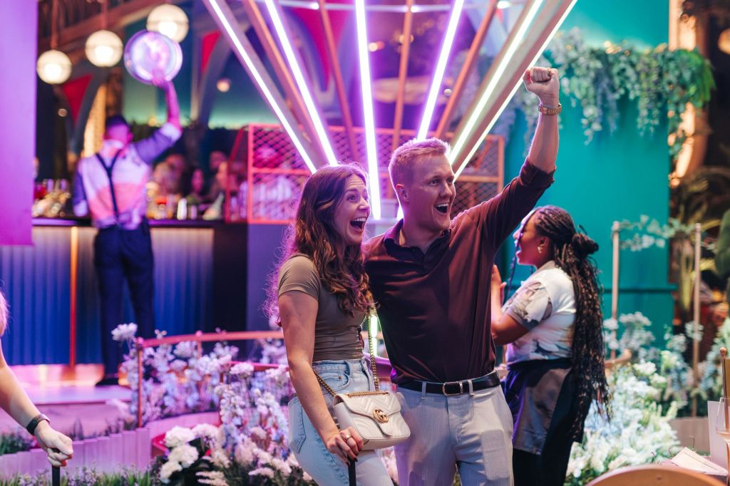 화려한 네온 불빛 앞에서 한 커플이 환호하며 미소를 짓고, 다른 한 여성이 배경에 서 있습니다.