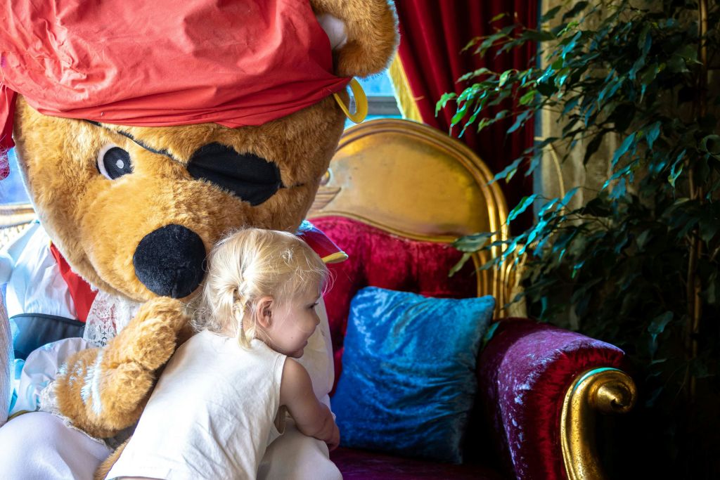 근처에 장식용 베개와 잎이 무성한 식물이 있는 레드 벨벳 의자에 앉아 곰 의상을 입은 사람을 껴안고 있는 아이.