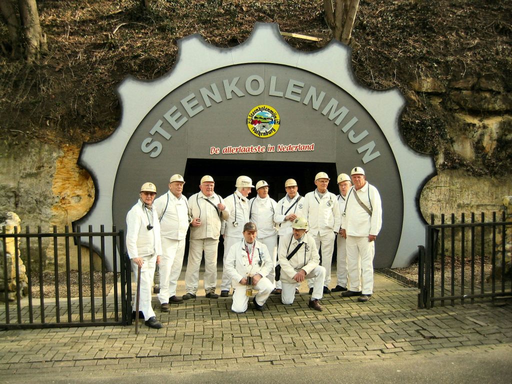 흰 유니폼과 헬멧을 입은 사람들이 "STEENKOLENMIJN" 표지판이 있는 광산 입구 앞에 서 있습니다.