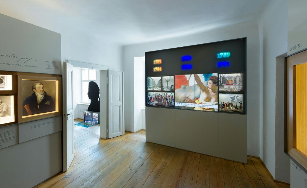 벽에 액자로 된 초상화, 다른 방으로 이어지는 열린 문, 패널에 비춰진 사진과 텍스트가 있는 박물관 방.