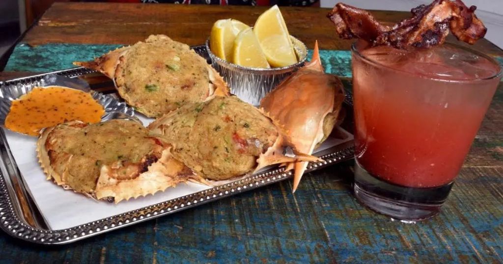 베이컨으로 장식한 레드 칵테일 한 잔에 게, 레몬 웨지, 속을 채운 소품을 나무 테이블에 올려 서빙합니다.