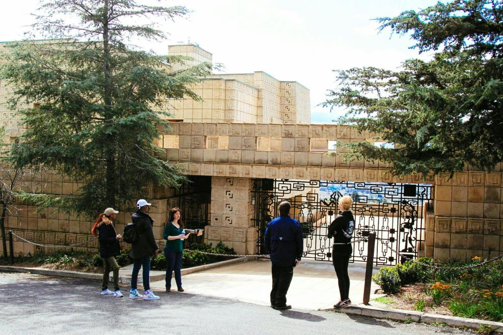 나무로 둘러싸인 기하학적 석조 건물의 문 근처에 한 무리의 사람들이 서 있습니다.