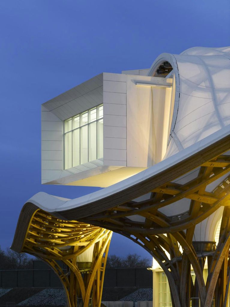 나무와 천으로 만든 곡선형 지붕에서 황혼빛 하늘을 배경으로 커다란 조명 흰색 상자 구조가 있는 현대적인 건물입니다.