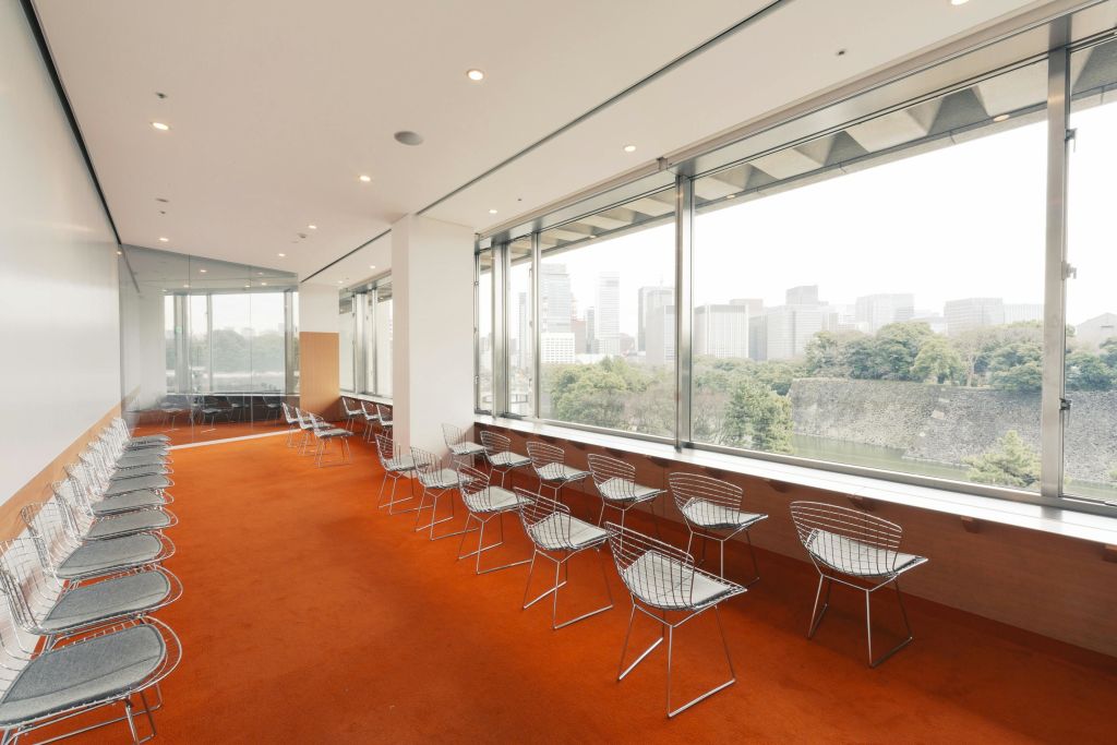 주황색 카펫, 금속 와이어 의자, 커다란 창문이 있는 객실로 나무와 건물이 있는 도시 풍경을 감상할 수 있습니다.