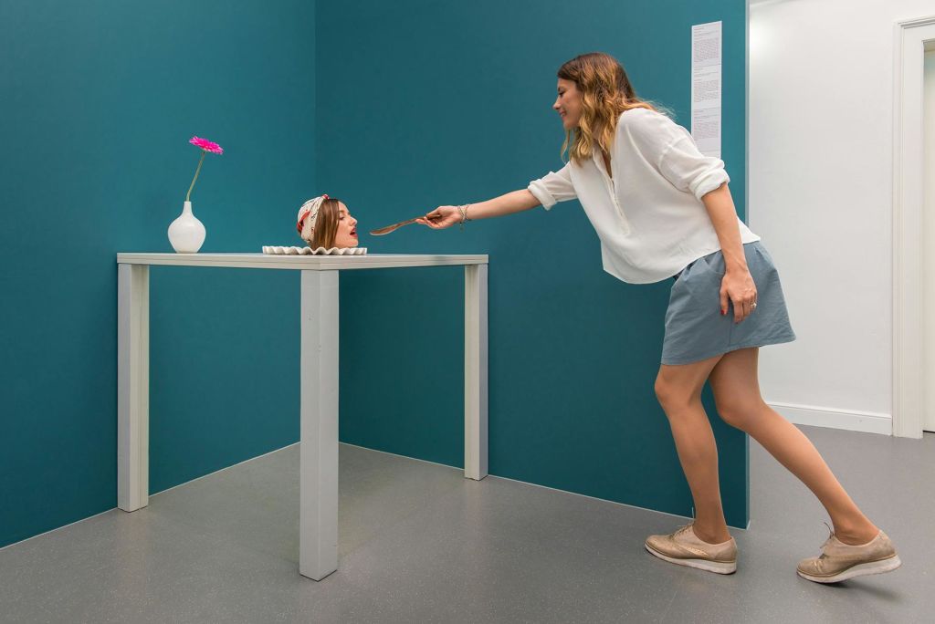 한 여성이 청록색 벽으로 둘러싸인 방의 탁자 위에서 몸이 벗겨진 머리를 숟가락으로 먹이는 척 장난스럽게 행동합니다.