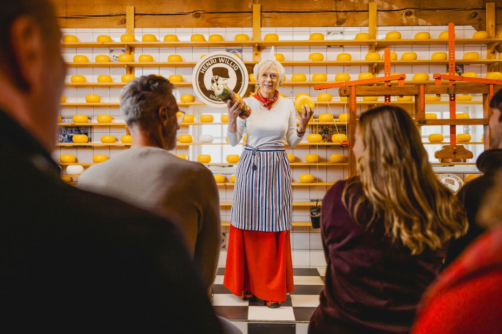 앞치마를 입은 한 여성이 치즈 바퀴가 채워진 선반 앞에 서서 관객에게 치즈를 들고 보여줍니다.