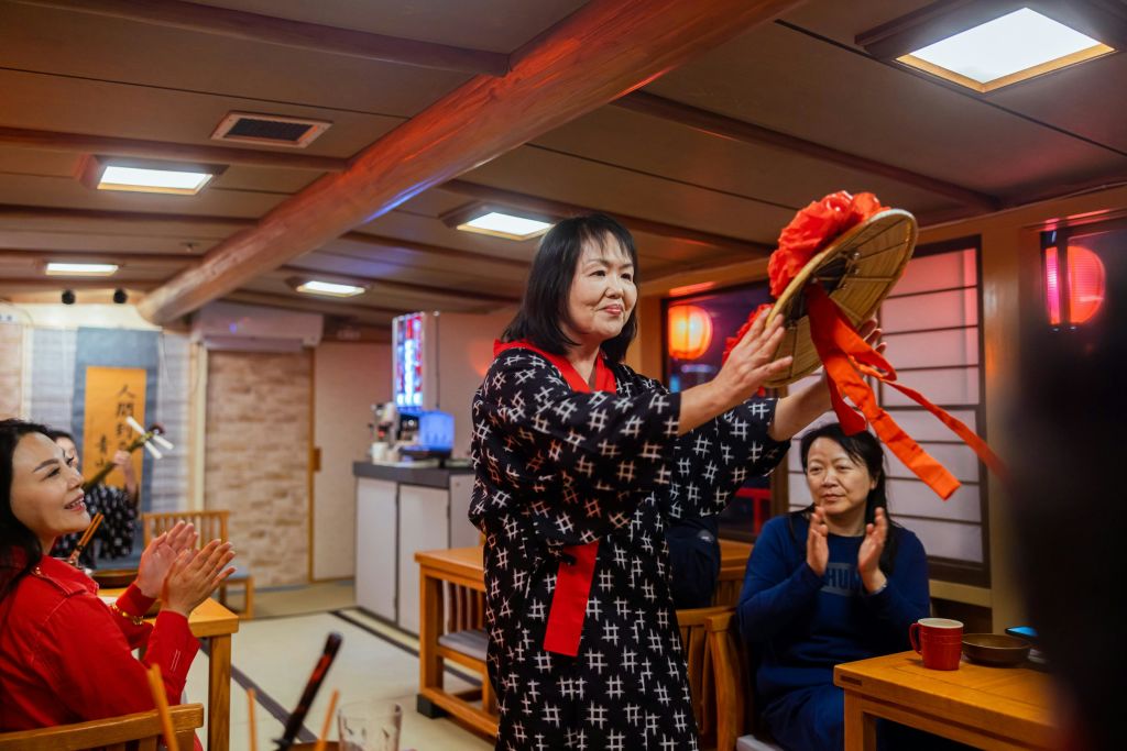 전통 복장을 한 여성이 방에서 공연을 펼치고 앉아 있는 사람들이 박수를 치고 있습니다. 객실은 낮은 조명과 일본식 요소로 꾸며져 있습니다.