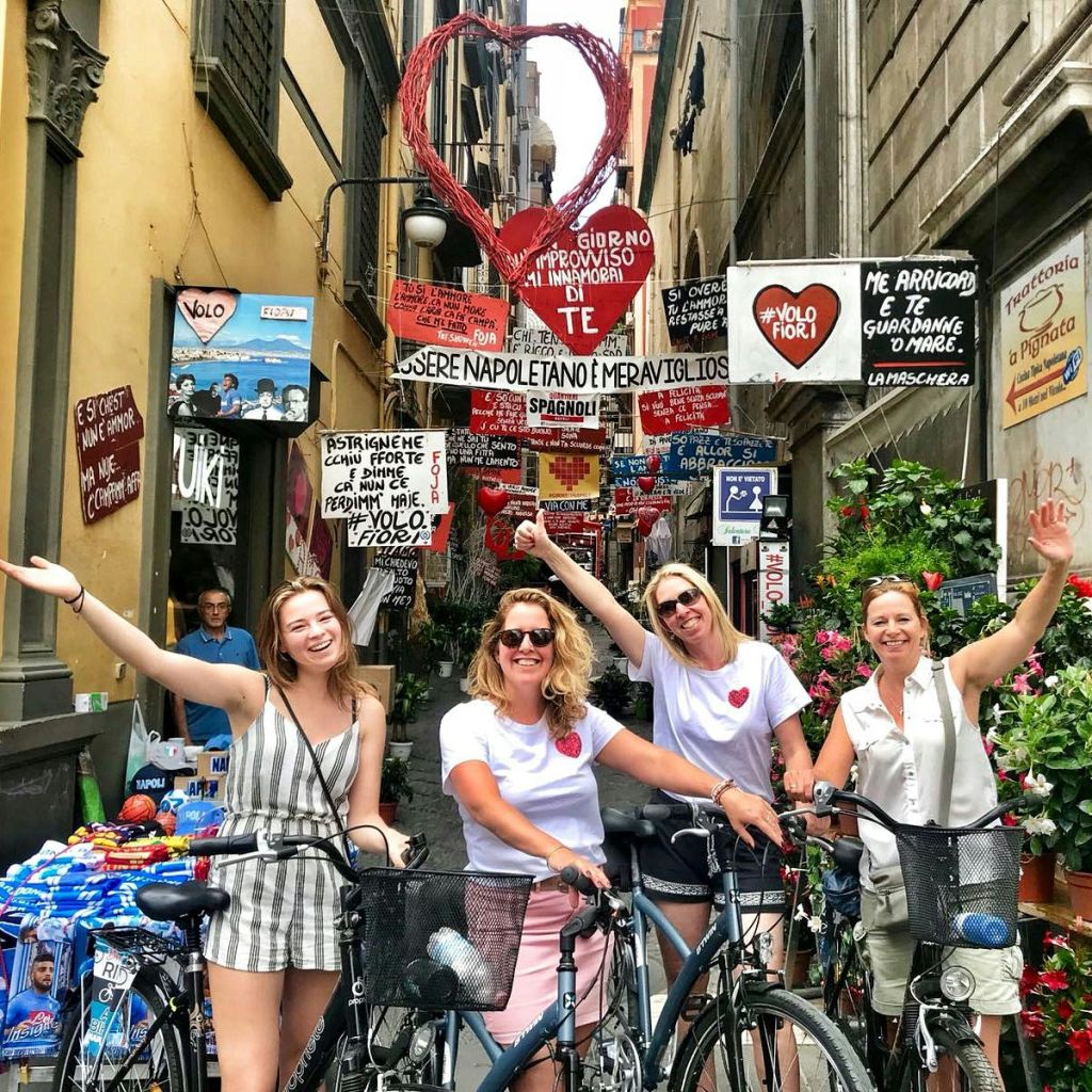 네 명의 여성이 간판과 장식으로 가득한 좁고 화려한 골목에서 자전거와 함께 서서 미소를 짓고 손을 흔들고 있습니다.