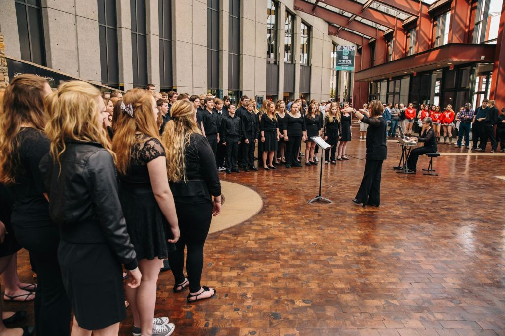 검은색 옷을 입은 대형 합창단이 커다란 창문이 있는 현대식 건물 앞 나무 바닥에서 실내 공연을 펼치고 있습니다.