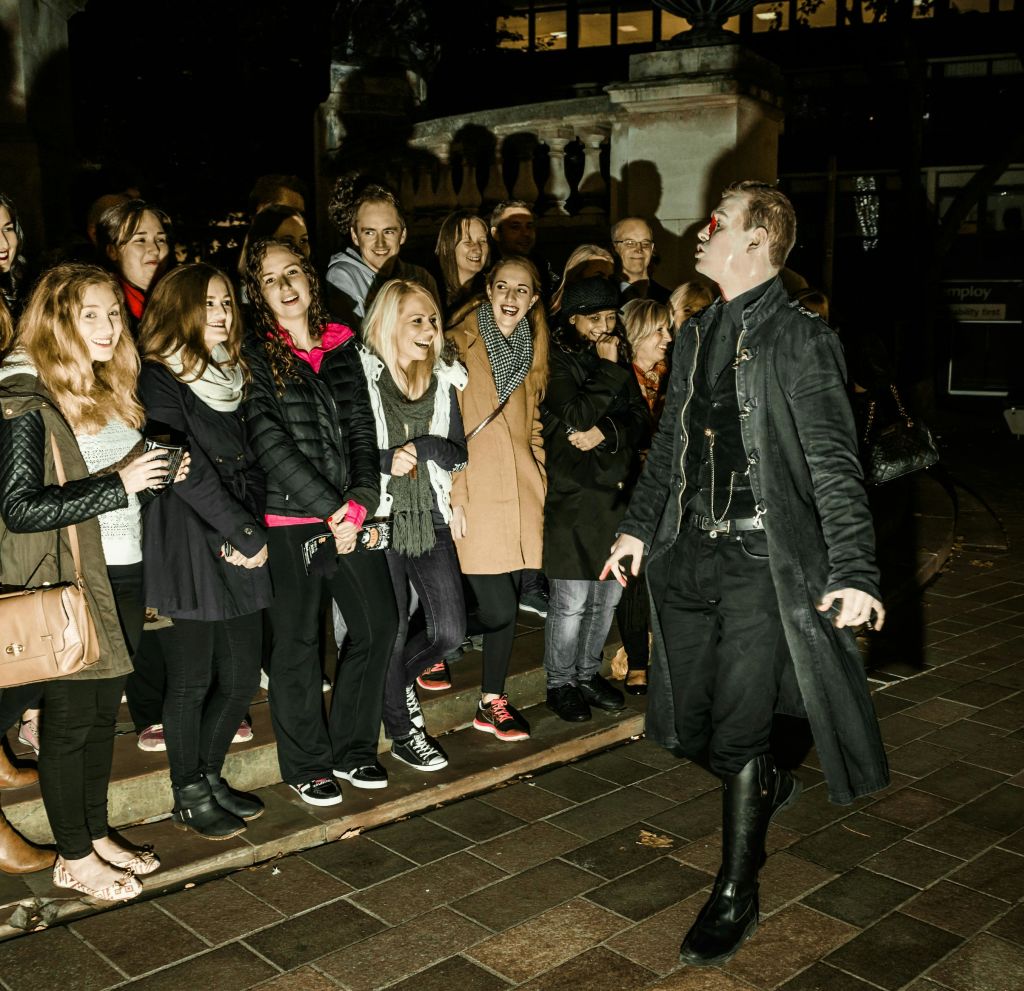 한 무리의 사람들이 길가에 서서 웃고 있습니다. 길고 어두운 코트를 입은 애니메이션 인물을 보면서 웃고 있습니다.