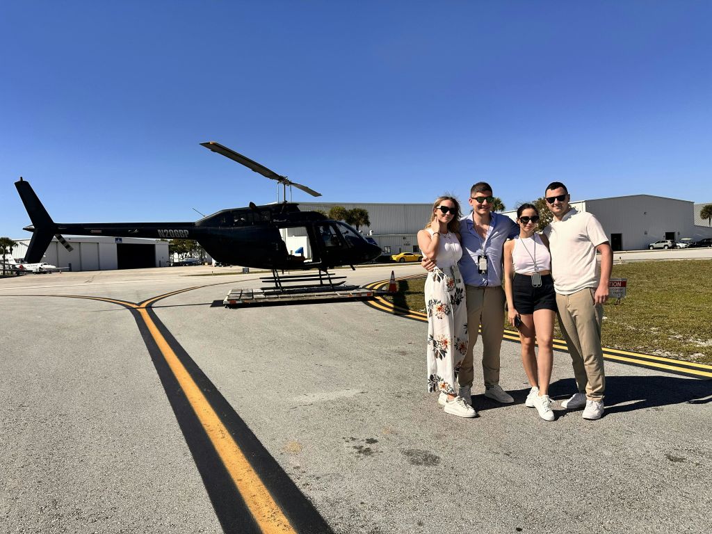 맑고 푸른 하늘 아래 활주로에 주차된 검은색 헬리콥터 앞에 서 있는 네 사람.