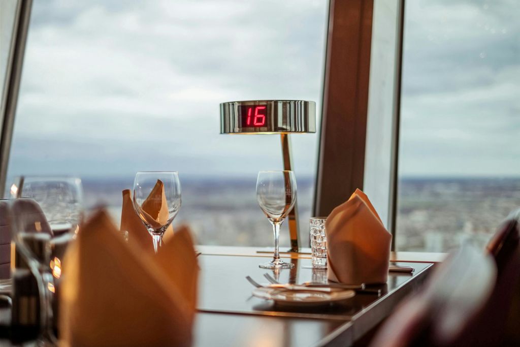 유리잔, 식기류, 냅킨이 놓인 레스토랑 테이블로, 도시 풍경이 내려다보이는 커다란 창문 앞에 테이블 번호 '16' 이 표시되어 있습니다.