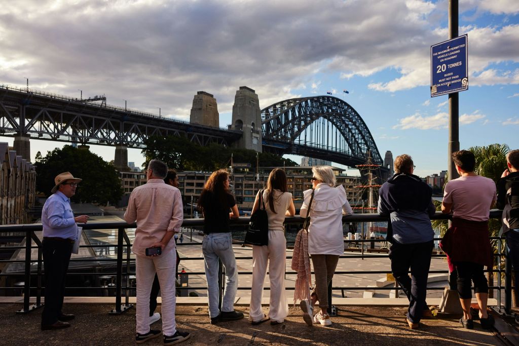 한 무리의 사람들이 수변 난간에 서서 부분적으로 흐린 하늘 아래 시드니 하버 브리지와 마주보고 있습니다.