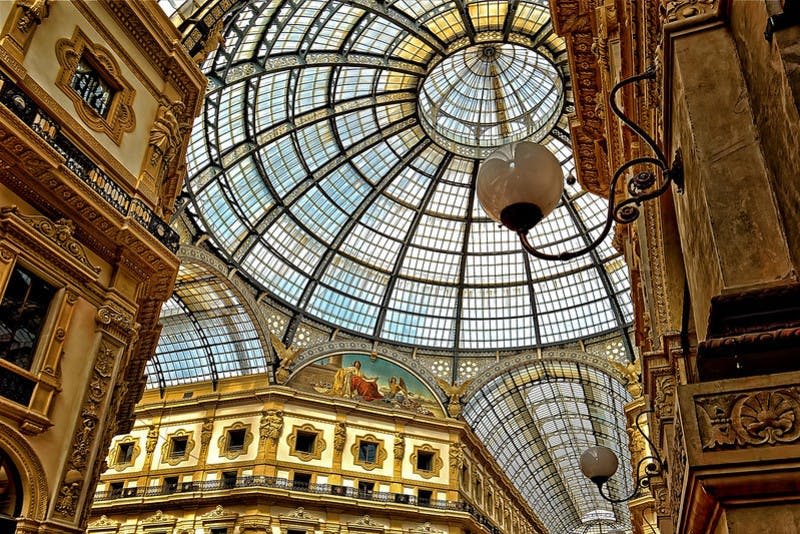 웅장하고 유서 깊은 건물 내부에 정교한 금속 프레임워크가 있는 화려한 유리 돔형 천장. 샹들리에와 장식 디테일이 보입니다.