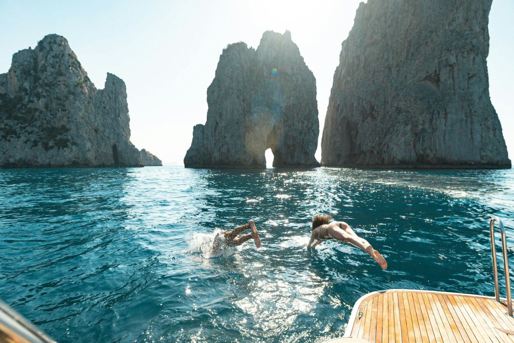 햇살이 내리쬐는 하늘 아래 커다란 암석 지층 근처에서 배를 타고 맑고 푸른 바다로 뛰어드는 두 사람.