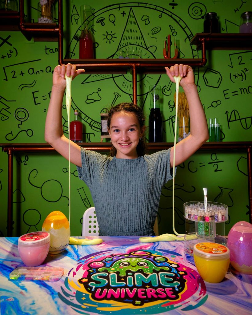 양손으로 슬라임을 뻗으며 웃고 있는 소녀. 그녀는 과학을 주제로 한 배경으로 점액 항아리가 있는 테이블에 앉아 있습니다.