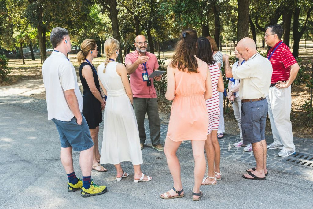 한 무리의 사람들이 공원에서 야외에 서서 가이드가 태블릿을 들고 말하는 것을 듣고 제스처를 취하고 있습니다.