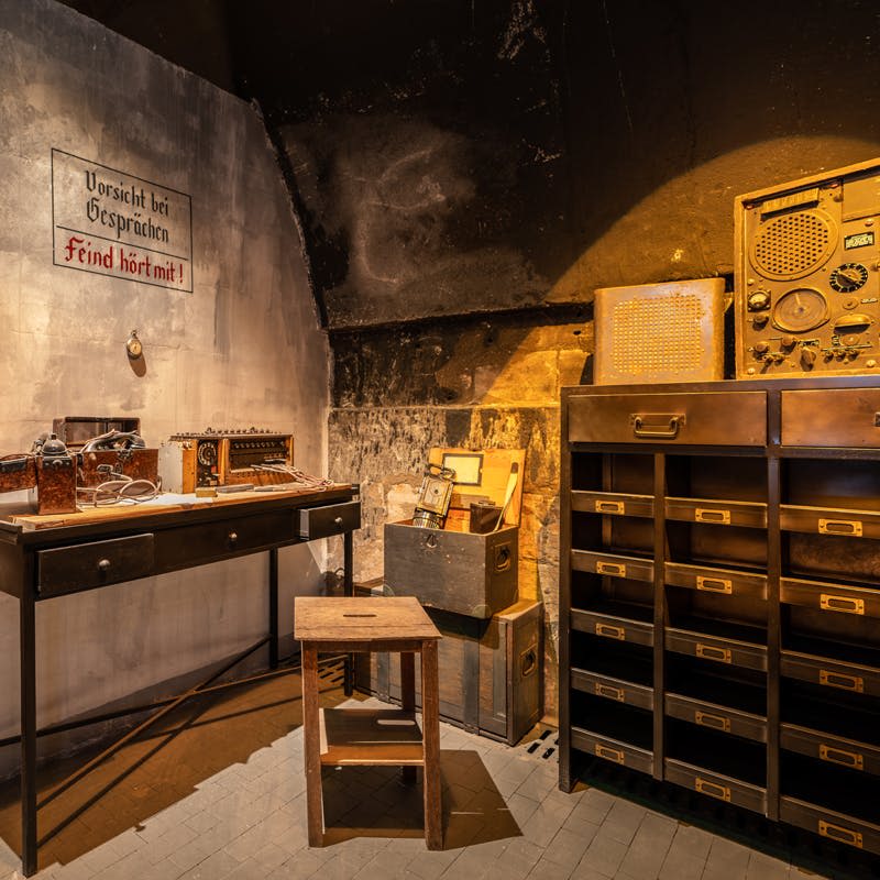 책상 위에 빈티지 라디오 장비, 의자, 수납함, 라벨이 붙은 서랍이 있는 캐비닛이 있는 조명이 희미한 객실입니다.