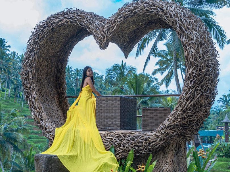 노란색 원피스를 입은 여성이 커다란 하트 모양의 고리버들 구조물 안에서 야자수와 흐린 하늘을 배경으로 포즈를 취하고 있습니다.