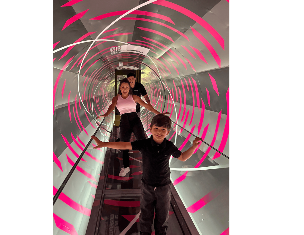 분홍색과 흰색 줄무늬가 있는 회전하는 터널을 걷는 세 사람이 균형을 위해 난간을 잡고 있습니다.