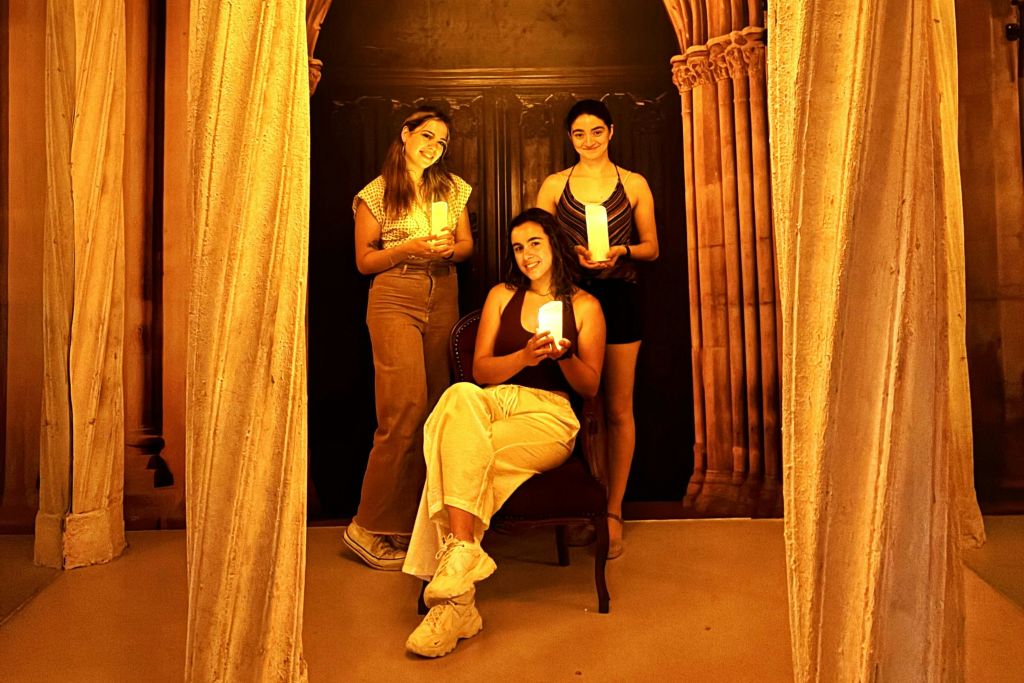 촛불을 든 세 사람이 장식 기둥과 어두운 화려한 배경으로 따뜻하게 빛나는 분위기에서 포즈를 취하고 있습니다.