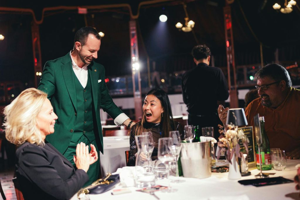 식탁에 앉아 초록색 정장을 입은 서 있는 남성을 보며 미소를 짓고 있는 여성. 다른 손님들과 빈 안경이 보입니다.