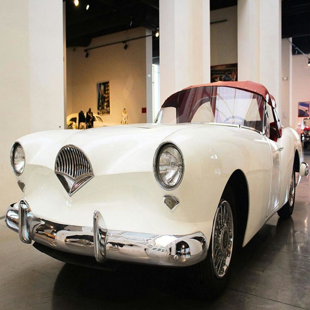 크롬 디테일과 빨간색 인테리어가 돋보이는 빈티지한 흰색 컨버터블 자동차가 실내 쇼룸에 주차되어 있습니다.