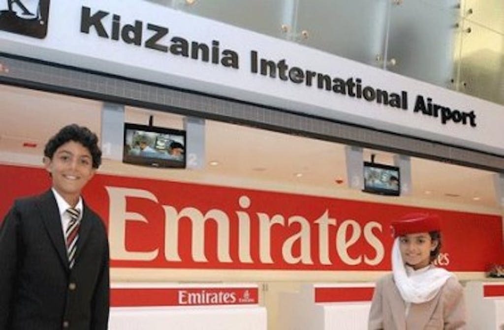 수트를 입은 미소 짓는 소년이 유명한 에미레이트 항공 브랜드가 있는 키자니아 국제 지역 앞에 서 있습니다.