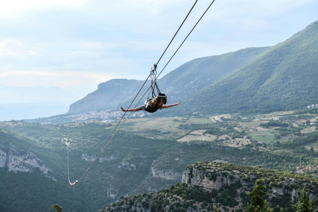 부분적으로 흐린 하늘 아래 계곡과 녹지가 있는 경치 좋은 산악 풍경 위에 사람이 집라인을 타고 있습니다.