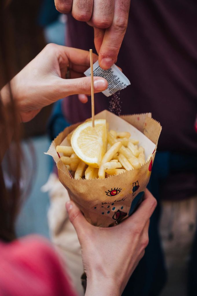 한 사람이 작은 나무 막대기를 사용하여 레몬 웨지를 얹은 감자 튀김 콘에 양념을 뿌리고 있습니다.
