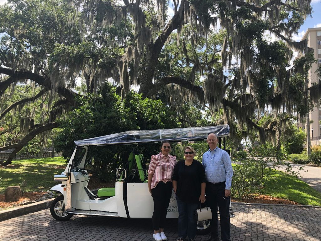 녹색 좌석이 있는 흰색 툭툭 차량 앞에 세 사람이 서 있으며, 커다란 나뭇가지 아래에는 스페인의 이끼가 덮여 있습니다.