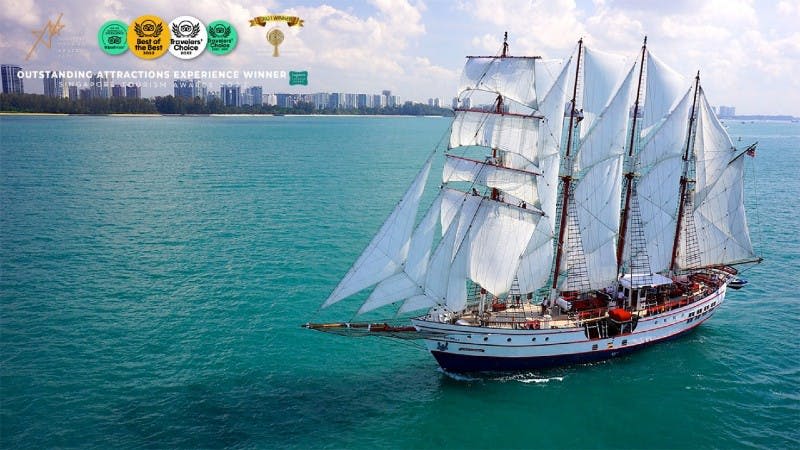 부분적으로 흐린 하늘 아래 도시 스카이라인 근처의 맑은 청록색 바다에서 하얀 돛이 달린 키 큰 배.