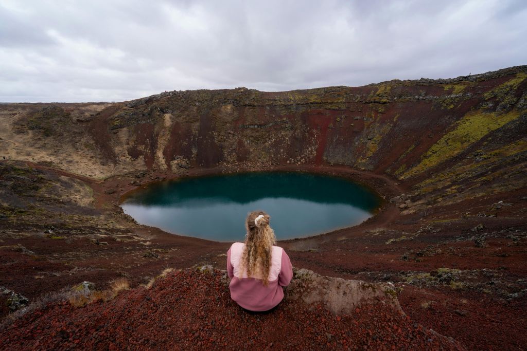 한 여성이 가을에 분화구가 내려다보이는 곳에 앉아 있습니다. 붉은 바위와 초록 이끼