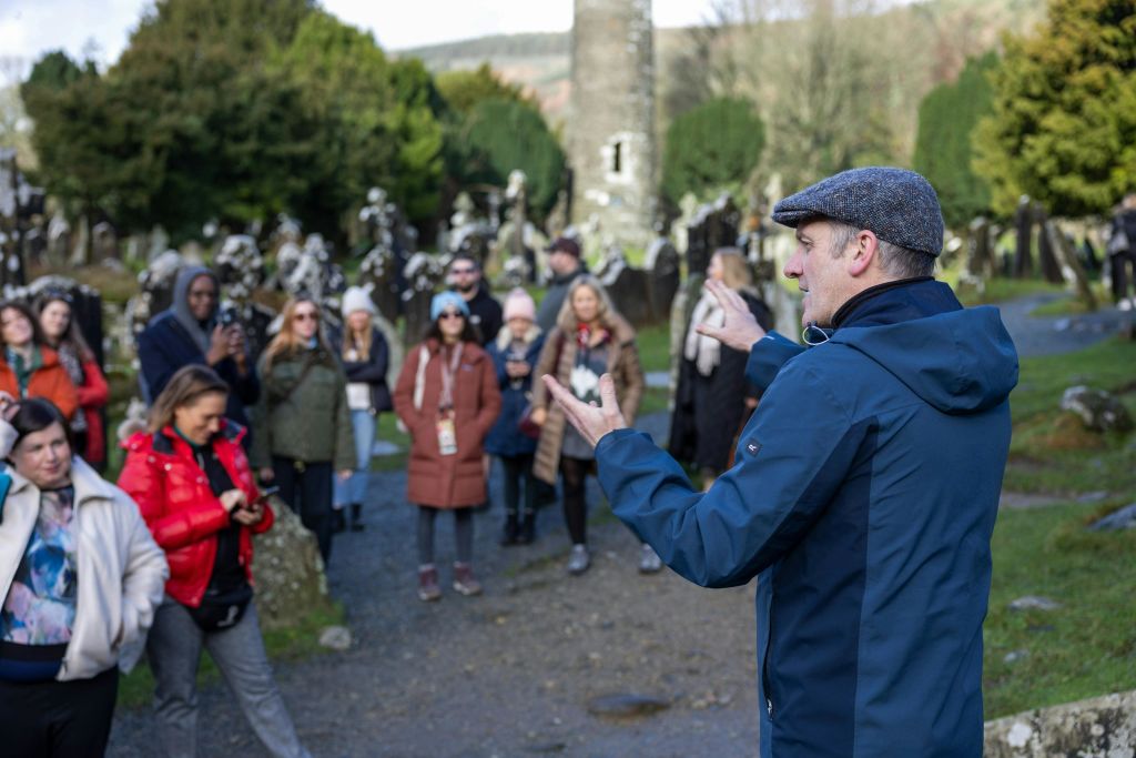 묘비와 탑을 배경으로 야외에서 일행과 이야기를 나누는 투어 가이드.