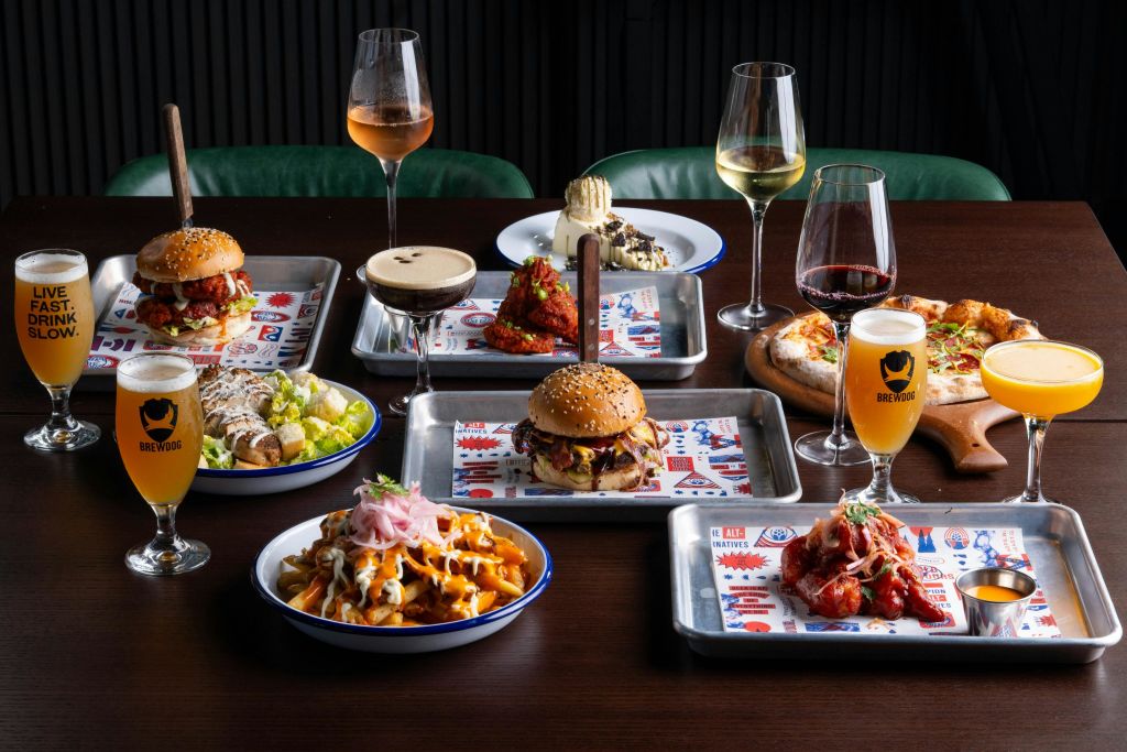 나무 테이블에 다양한 햄버거, 파스타, 음료가 놓여 있으며, 와인, 맥주, 칵테일도 함께 제공됩니다.