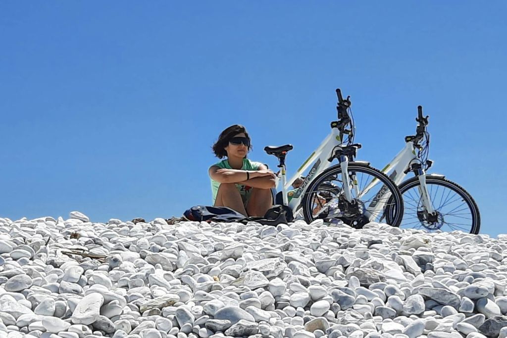 선글라스를 착용한 사람이 맑고 푸른 하늘 아래 자전거 2대 옆의 바위 해변에 앉아 있습니다.