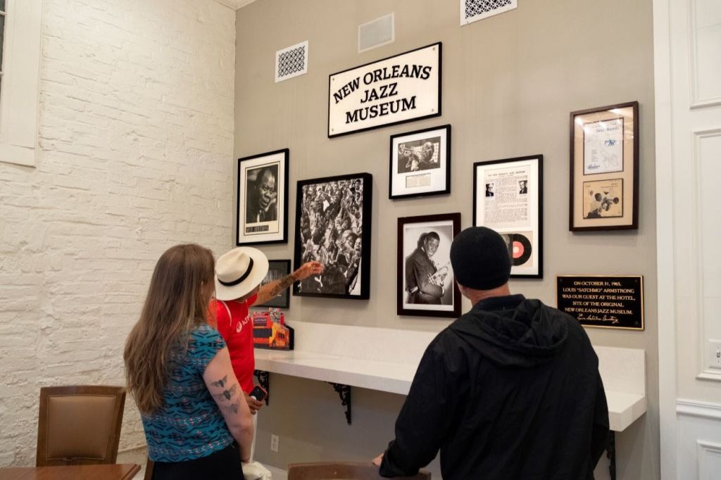 세 사람이 박물관 벽에 걸린 액자에 담긴 재즈 기념품을 살펴보고, 그 위에 '뉴올리언스 재즈 박물관' 이라는 간판이 있습니다.