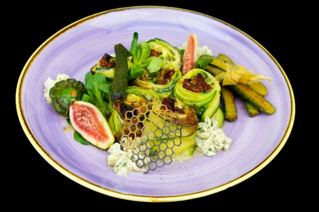 보라색 접시에 애호박 롤, 무화과, 채소, 장식용 벌집 격자를 담은 다채로운 요리입니다.