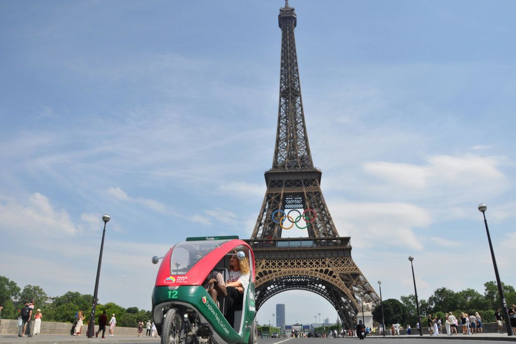 에펠탑 앞에 승객이 타고 있는 3륜 페디캡으로, 구조물에는 올림픽 링이 표시되어 있습니다.