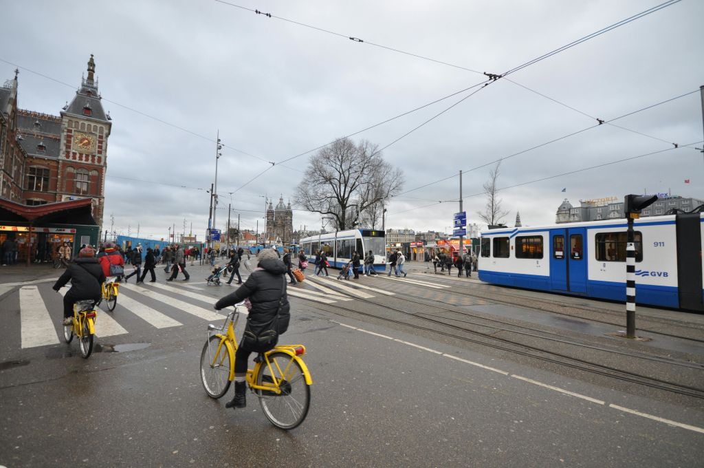 노란색 자전거를 타는 사람이 흐린 하늘 아래 보행자, 트롤리, 머리 위 전선이 있는 바쁜 거리를 가로지르고 있습니다.