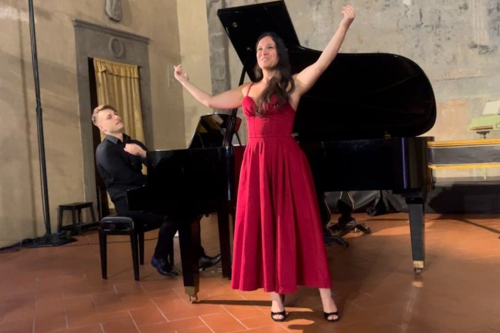 빨간색 드레스를 입은 여성이 그랜드 피아노 옆에 팔을 들고 당당하게 서 있고, 검은 양복을 입은 남성이 피아노를 연주합니다.