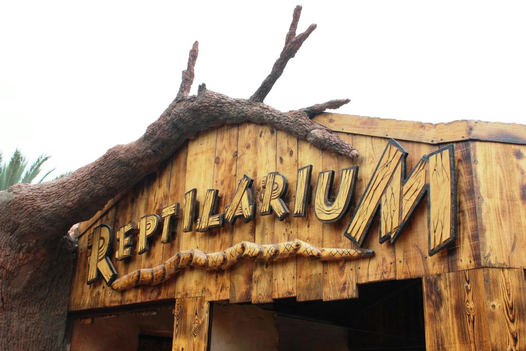 "Reptilarium" 표지판이 있는 목조 건물 외관에는 가짜 나뭇가지와 뱀 모형이 있습니다.