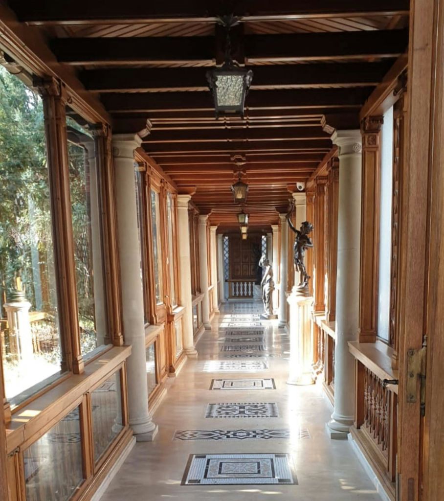 왼쪽에는 유리창, 조각상, 기둥, 무늬가 있는 바닥 타일이 있는 나무 복도가 문으로 이어집니다.