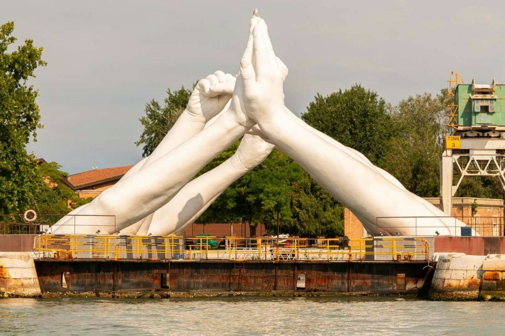 푸르름을 배경으로 운하를 배경으로 기도하는 듯한 몸짓으로 손을 모은 커다란 흰색 조각상.