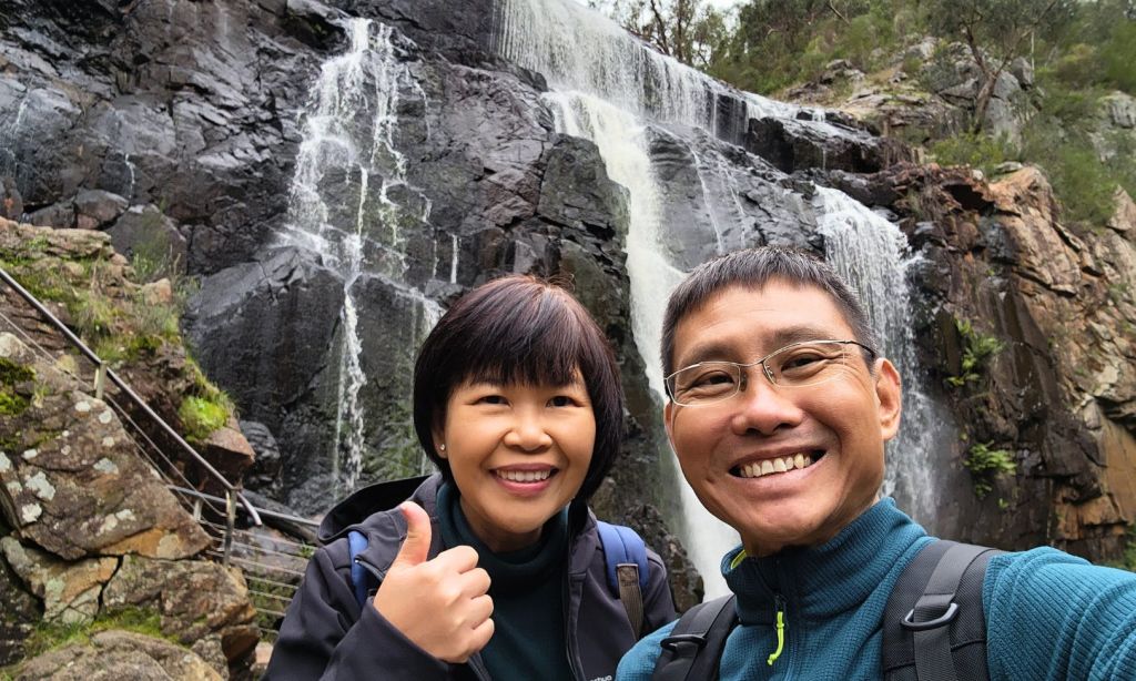 바위 지형과 녹지를 배경으로 폭포 앞에서 미소 짓는 두 사람이 포즈를 취하고 있습니다.