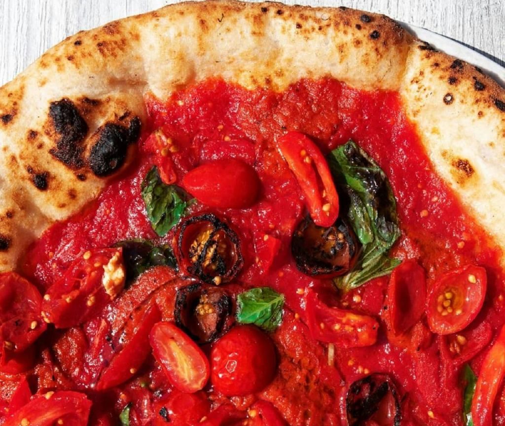 토마토 소스, 체리 토마토, 구운 채소, 신선한 바질이 토핑된 숯불 크러스트 피자의 클로즈업.