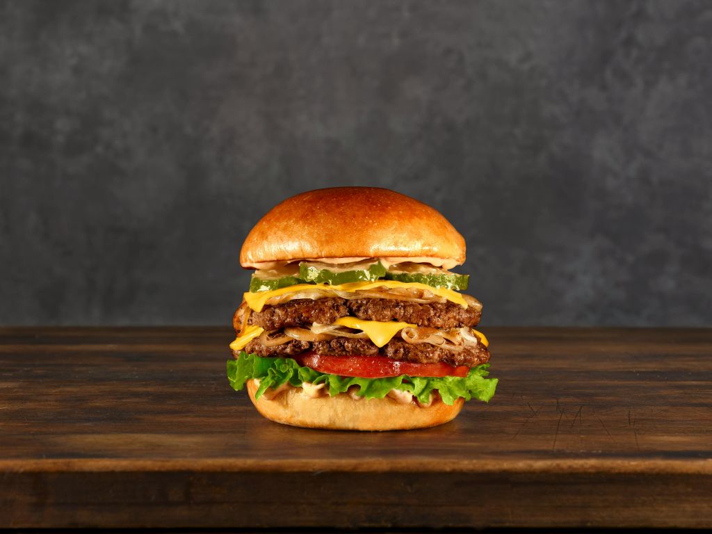 어두운 배경에 나무 표면에 양상추, 토마토, 피클, 치즈를 올린 더블 치즈버거입니다.